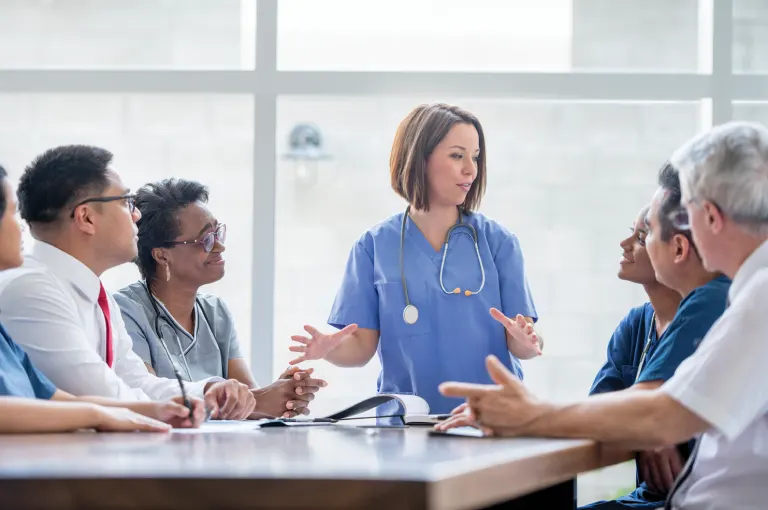 Actor portrayals of female nurse in blue scrubs with short, dark brown hair and a stethoscope around her neck talking to other male and female medical staff.